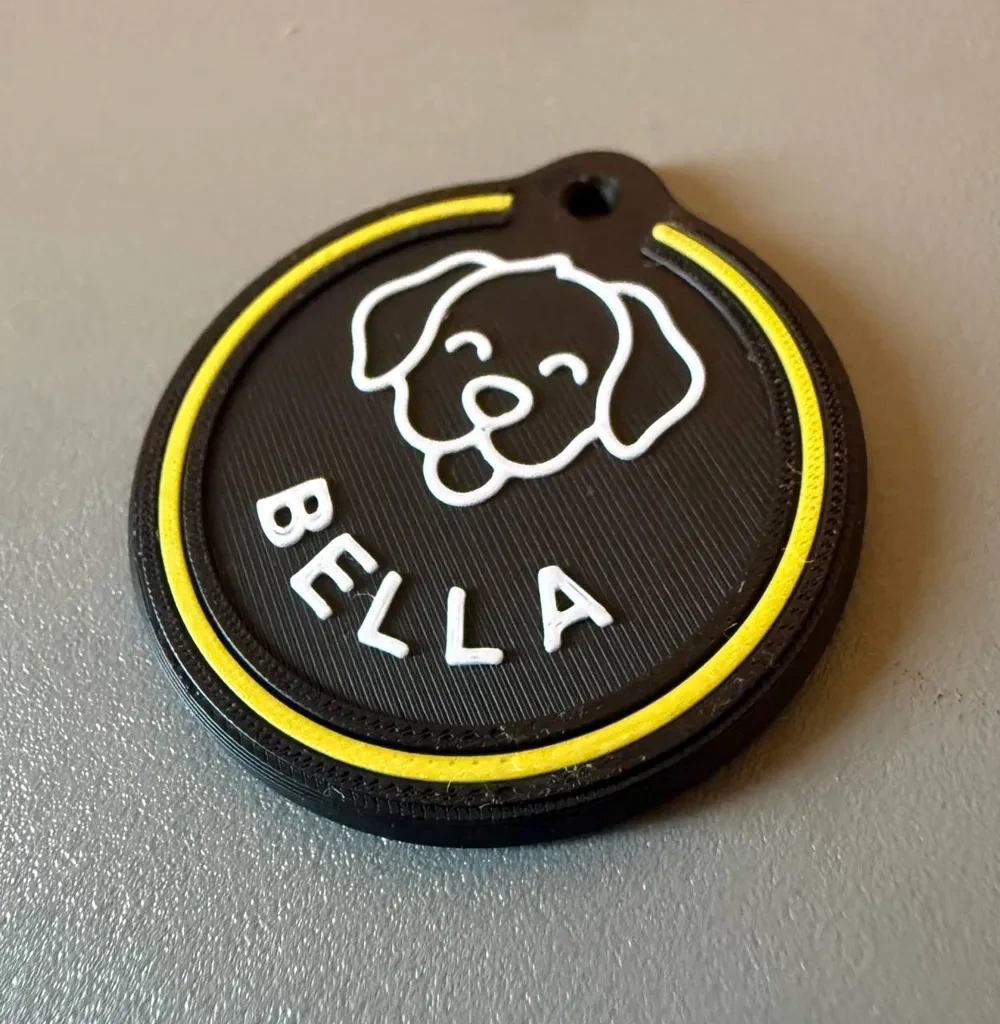 A round black pet ID tag with a white dog illustration, BELLA in white letters and a yellow outline.